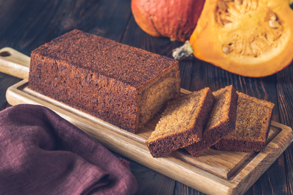 A freshly baked pumpkin banana loaf sliced on a wooden cutting board with a halved pumpkin in the background.