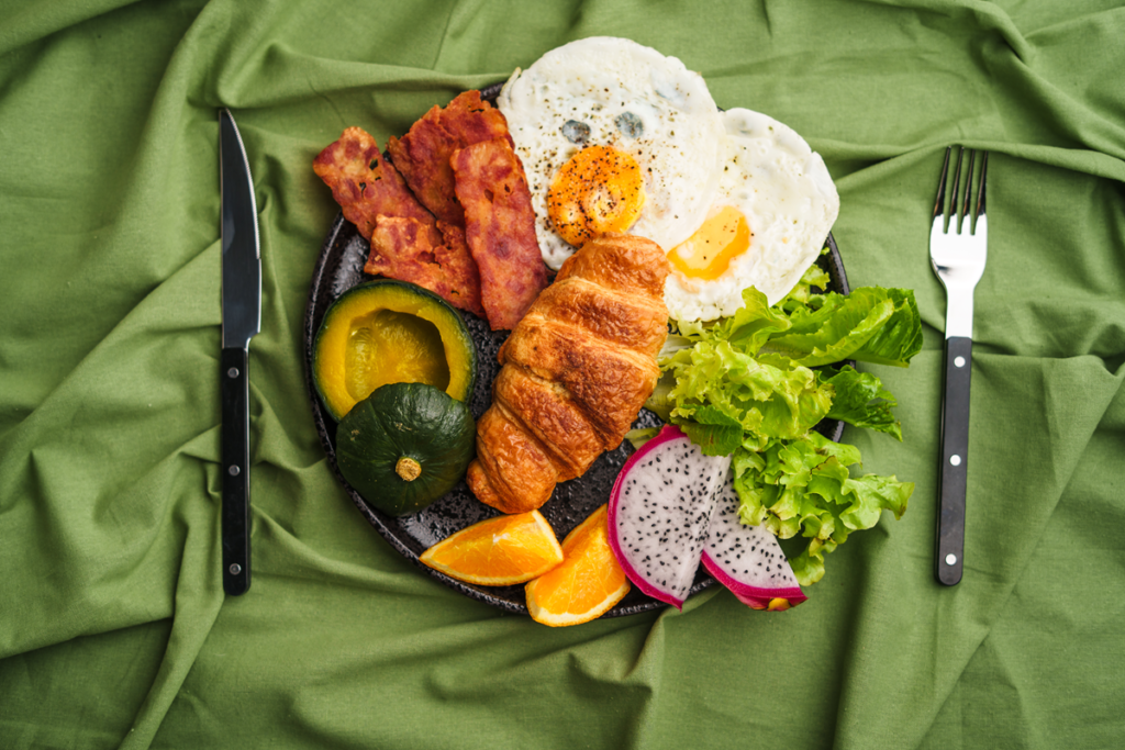 Healthy breakfast plate with croissant, fried eggs, bacon, and fresh vegetables