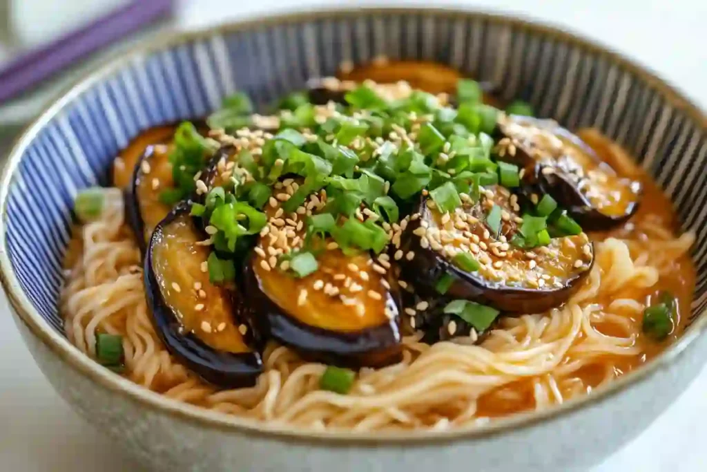 A bowl of miso noodles with tender eggplant and shiitake mushrooms, garnished with green onions and sesame seeds.