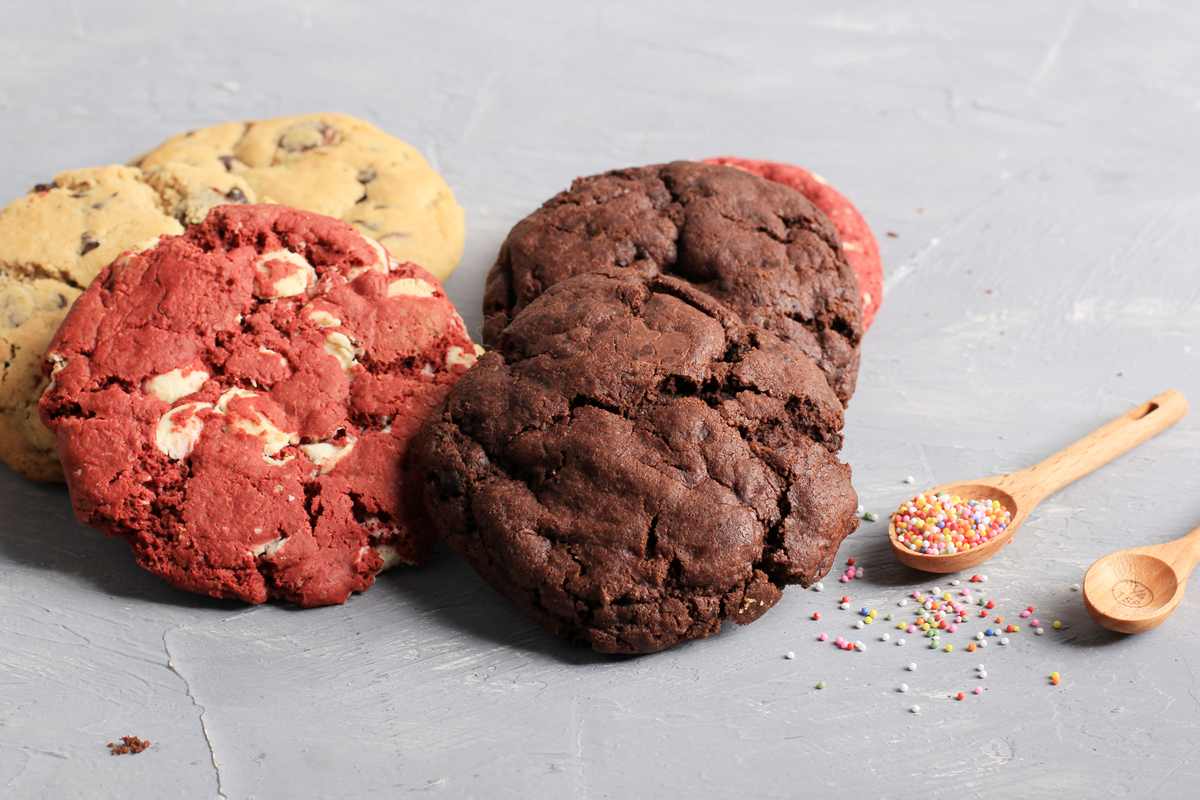 Various soft American cookies, including vanilla, red velvet, and chocolate, displayed on a gray background with colorful sprinkles.