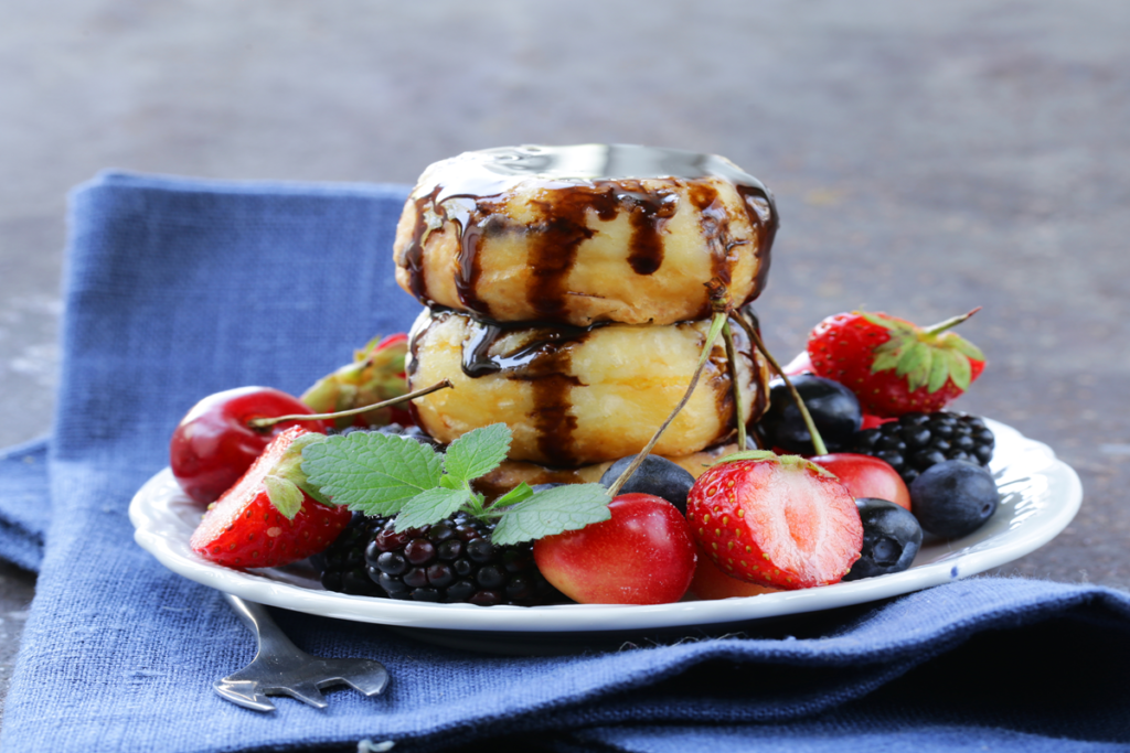 Two stacked fluffy cottage cheese pancakes drizzled with chocolate syrup, served with fresh berries like strawberries, blackberries, and cherries on a plate.