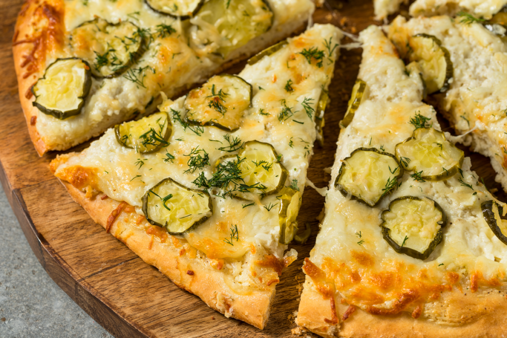 Close-up of sliced dill pickle pizza with cheese and fresh dill on a wooden serving board.